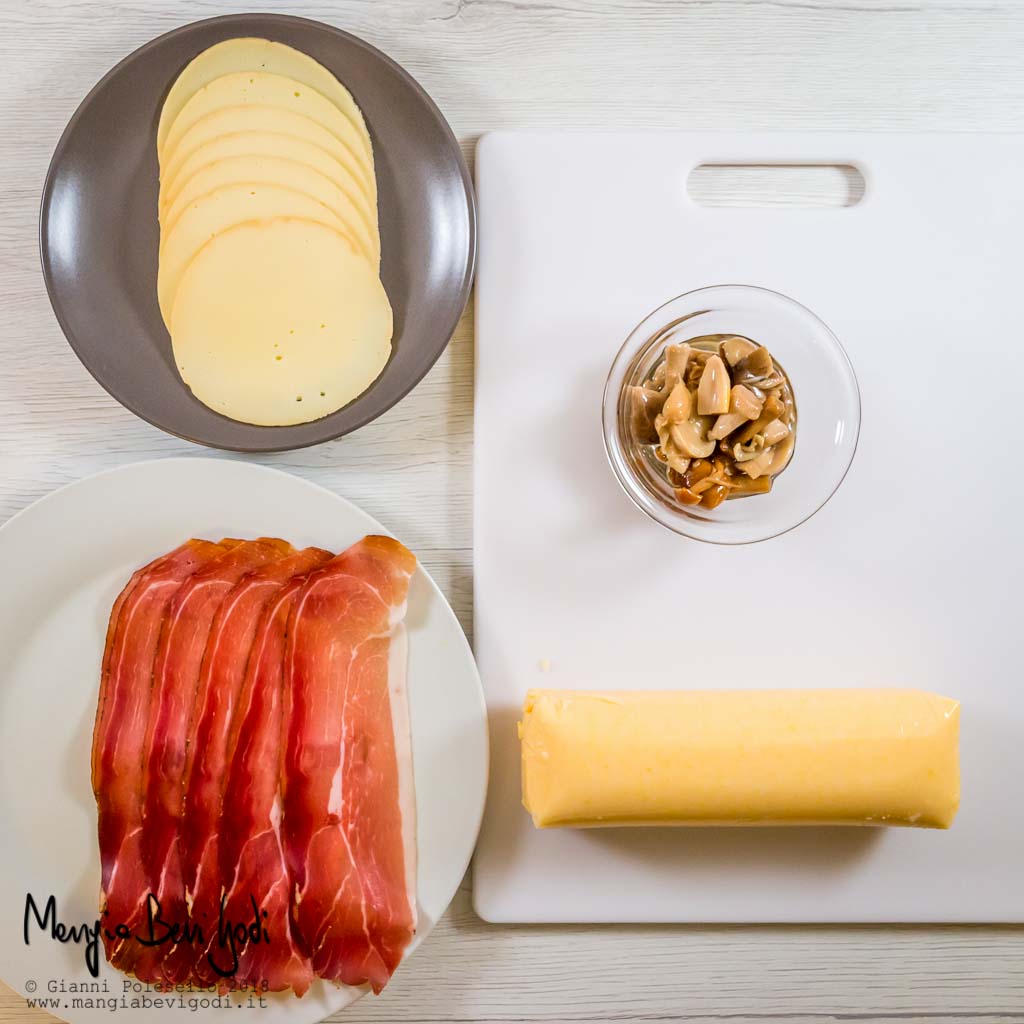 Gli ingredienti: polenta gialla pronta sopra tagliere bianco, ciotola di vetro con funghi sott'olio, piatto bianco con fettine di speck, piatto marrone con fettine di provola