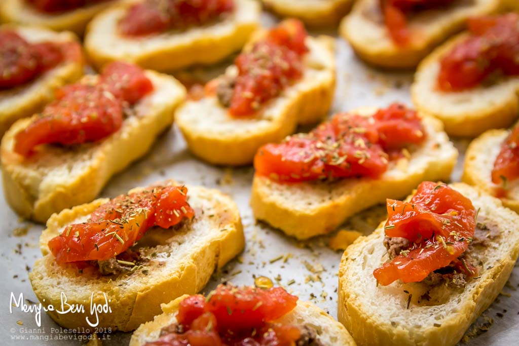 Crostini con pasta d'acciughe e pelati, spolverati da origano secco su placca rivestita di carta da forno